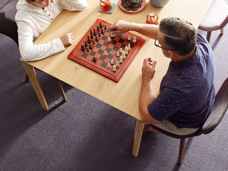 two men playing chess at a wooden table in a room with purple carpet from Northcraft Flooring & Design in Raytown, MO